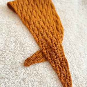 May include: Mustard-colored knitted head covering with a textured, diamond-patterned design. The head covering is made of a soft, warm material and is likely handmade. The item is laying on a white, textured surface.