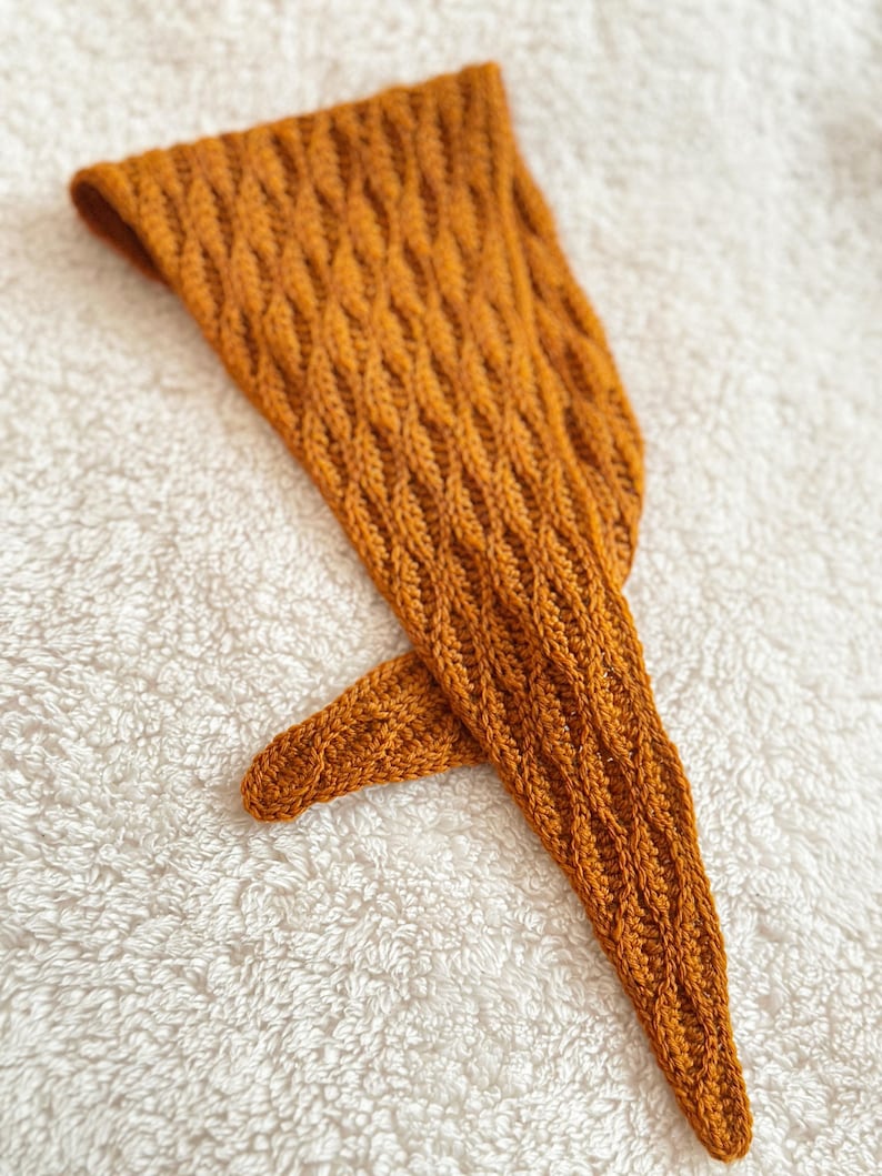 May include: Mustard-coloured knitted head covering with a textured, diamond-patterned design. The head covering is made of a soft, warm material and is likely handmade. The item is laying on a white, textured surface.