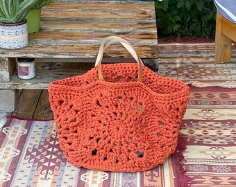 May include: A large, orange crocheted tote bag with a wooden handle. The bag has a circular pattern and is sitting on a patterned rug.