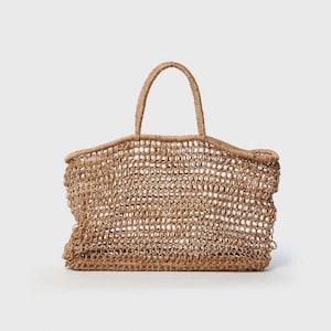 Puede incluir: Un bolso tote tejido en color marrón natural. El bolso tiene forma rectangular con una abertura ancha y dos asas. El diseño de tejido abierto es visible, creando una apariencia texturizada.