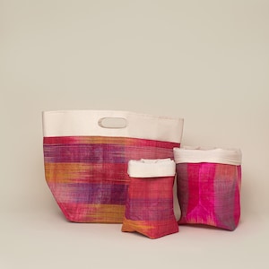 May include: Three woven fabric storage bags in varying sizes. The bags feature horizontal stripes in shades of pink, orange, and purple. Each bag has a cream-colored top and a handle on the largest bag.