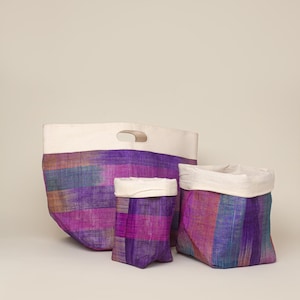 May include: Three woven storage baskets in varying sizes. The baskets feature a striped pattern in shades of purple, pink, and blue, with a cream-colored top. The largest basket has a handle.