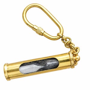 May include: A gold-toned metal keychain featuring a cylindrical casing with a clear window revealing a small hourglass inside. The keychain includes a sturdy oval-shaped carabiner clip attached by a short metal chain to the end of the cylinder.