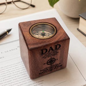 Puede incluir: Un bloque de madera cuadrado de color marrón oscuro con una brújula circular de latón incrustada en la parte superior. La cara frontal está grabada con la palabra DAD, una rosa de los vientos y el texto: MY FIRST HERO. MY FOREVER GUIDE.