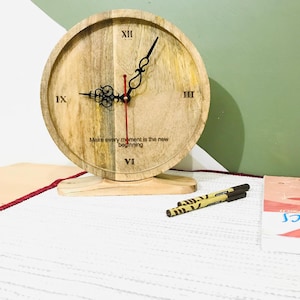 May include: A circular wooden clock with Roman numerals and ornate black hands. The clock face features the text "Make every moment is the new beginning". The clock sits on a wooden stand. Two pens are in the foreground.