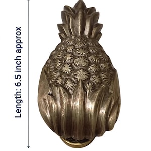 May include: A brass pineapple door knocker with an antique finish. The text BRASS PINEAPPLE DOOR KNOCKER — DIMENSIONS is at the top. The image indicates a length of 16.51 cm and a width of 7.62 cm. The material is solid brass with a screw-on back plate.
