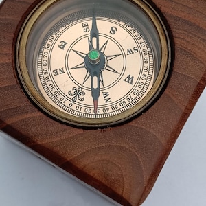Puede incluir: Un bloque de madera cuadrado que alberga una brújula circular de latón. La esfera de la brújula presenta un fondo dorado metálico con puntos cardinales negros N, E, S, O, SE, SO, NO y NE, junto con una escala de grados y una aguja central.