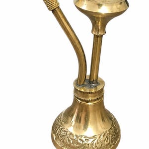 May include: A polished brass smoking pipe featuring a wide, circular bowl and a curved stem with a textured, diamond-patterned grip. The base of the pipe is rounded and decorated with an intricate, embossed floral relief pattern around the lower edge.