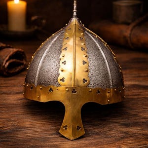 Puede incluir: Un casco de estilo medieval con acabado plateado y dorado. El casco presenta una parte superior redondeada con una punta central y una banda dorada decorativa alrededor de la base. El diseño incluye remaches y una distintiva protección facial.