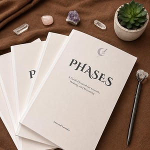 Könnte beinhalten: Eine Ansammlung weißer Journale mit dem Wort „PHASES“ auf dem Cover, zusammen mit dem Text „A Guided Journal for Growth, Healing, and Becoming“. Die Journale liegen auf einer braunen Oberfläche, zusammen mit Kristallen und einer kleinen Sukkulente. Ein Stift mit Kristallspitze ist ebenfalls vorhanden.