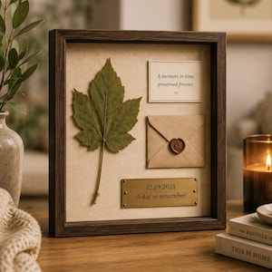 May include: A square dark wood shadow box frame containing a dried green maple leaf, a small envelope sealed with a brown wax seal, and a rectangular brass plaque engraved with the date 12.09.2023 and the text A day to remember.