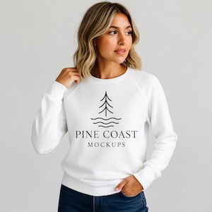以下が含まれることがあります： ラグランスリーブの白いスウェットシャツ。黒い松の木と水面の波のグラフィックがあり、その下に「PINE COAST MOCKUPS」の文字があります。スウェットシャツは人が着ています。