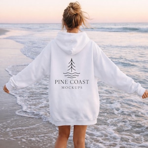 May include: White hooded sweatshirt featuring a black tree and wave graphic, with the text "PINE COAST MOCKUPS". This casual sweatshirt is ideal for everyday wear. The image is taken at the beach.