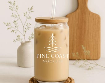 Op de afbeelding: Een heldere glazen beker gevuld met een lichtbruine drank en ijsblokjes, afgedekt met een houten deksel en een glazen rietje. De beker heeft een wit logo met de tekst "PINE COAST MOCKUPS". Het staat op een ronde, geweven onderzetter.