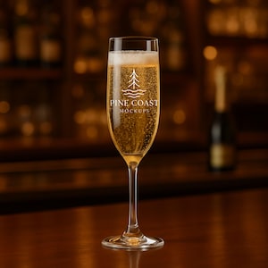 Puede incluir: Una copa de champán llena de un líquido dorado y burbujeante. La copa tiene un tallo largo y una copa ancha. El texto "PINE COAST MOCKUPS" está impreso en la copa. El fondo es una imagen borrosa de un bar.