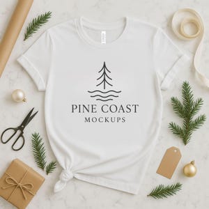 Op de afbeelding: Een wit t-shirt met de tekst "PINE COAST MOCKUPS" en een boomlogo. Het shirt is gestyled met vakantiethema decoraties, waaronder takjes groenblijvende planten, een gouden ornament en een geschenkdoos.