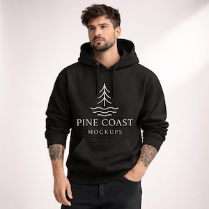 Peut inclure: Sweat à capuche noir avec un motif blanc d'arbre et de vagues, avec le texte "PINE COAST MOCKUPS". Le sweat-shirt a une poche avant et une capuche à cordon. Le modèle porte un jean noir.
