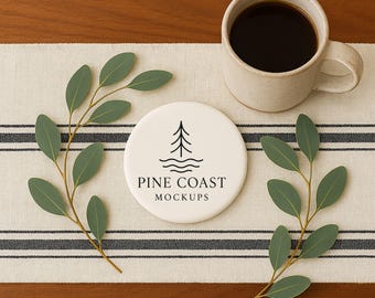 Puede incluir: Una taza de cerámica beige llena de café oscuro se encuentra sobre un corredor a rayas. Una posavasos blanco con un logotipo de árbol y el texto "PINE COAST MOCKUPS" está al lado de la taza. Ramas verdes de eucalipto a ambos lados.