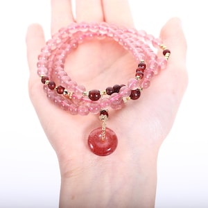 Pode incluir: Uma pulseira de várias voltas com contas de quartzo morango translúcido, intercaladas com contas esféricas vermelho-escuras e pequenos espaçadores de metal dourado. Um pingente de pedra circular, plano e vermelho-escuro, pende de um elo de metal dourado.