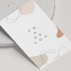 Puede incluir: Una tarjeta blanca con las palabras "THANK YOU" en una fuente minimalista, acompañada de formas abstractas en beige y marrón claro. La tarjeta está en ángulo sobre una superficie gris moteada.