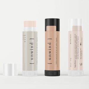 May include: Three lip balm tubes, one with a white label and the text "{ unwind }" in black, one with a beige label and the text "{ unwind }" in black, and one with a white label and the text "RITUALS" in black. The label on the beige tube reads "100% natural & for all skin types". The label on the white tube reads "3.4 oz - 3.3g" and lists the ingredients.