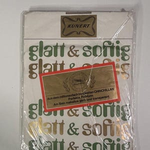 Pode incluir: Um pacote de meias KUNERT, embalado em plástico transparente. A embalagem é branca com as palavras "glatt & softig" em verde e dourado. Uma etiqueta dourada diz "CHINCHILLAN" e outro texto.