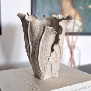 Könnte beinhalten: Eine strukturierte, hellgraue Keramikvase mit einem einzigartigen, gerüschten Design. Die Vase steht auf einem Buch mit dem Titel "Rembrandt Paintings & Portraits". Die Gesamtästhetik ist modern und künstlerisch.