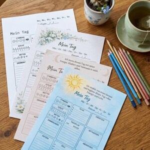 May include: A collection of "Mein Tag" daily planner sheets in various pastel colors, featuring floral and celestial designs. Colored pencils and a teacup with a tea bag are on a wooden surface.