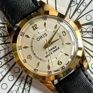 Peut inclure: Montre-bracelet Oris dorée avec un cadran crème et un bracelet en cuir noir. Le cadran affiche des aiguilles et des index dorés, avec le nom de la marque et le texte "17 Jewels Shock Proof". La montre est posée sur un fond à motifs.