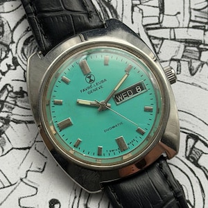 May include: A vintage Favre-Leuba Geneve Duomatic watch with a teal face and silver case. The watch features a black leather strap and displays the day of the week. The watch face has rectangular hour markers and the text "SWISS MADE".