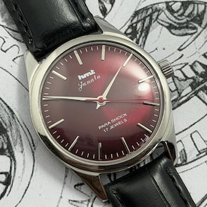 Peut inclure: Une montre-bracelet vintage avec un cadran bordeaux et des aiguilles argentées. Le cadran de la montre porte l'inscription "hmt Janala" et "Para Shock 17 Jewels". Elle possède un boîtier en acier inoxydable et un bracelet en cuir noir.