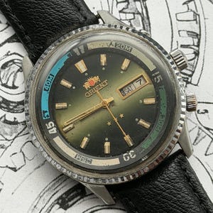 May include: Vintage Orient watch with a black leather strap. The watch face is dark green with gold hands and markers. The bezel has blue and green accents with white numerals. The watch displays the day and date.