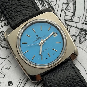 May include: A vintage Tudor Oyster-Prince-Junior automatic watch. It has a square stainless steel case and a light blue dial. The watch features gold hour markers and hands, a date window, and a black leather strap. The watch is Swiss made.