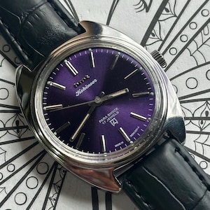 May include: A vintage wristwatch with a purple face and silver casing. The watch face features silver hour markers and the text "Kohinoor". The watch has a black leather strap.