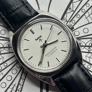 May include: Silver-toned wristwatch with a white face and black leather band. The watch face features black hour markers, black hands, and the text "PARA SHOCK 17 JEWELS". The watch has a square case and a black leather strap.