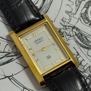 Peut inclure: Une montre rectangulaire Seiko Quartz avec un boîtier doré, un cadran blanc doté d'index dorés, les chiffres 12 et 6, et un bracelet en cuir noir à la finition texturée. Le cadran de la montre affiche le nom de la marque Seiko Quartz.