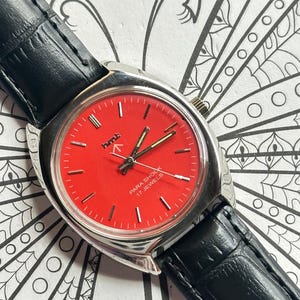 May include: A vintage wristwatch with a red face and silver casing. The watch has a black leather band and the text "PARA SHOCK 17 JEWELS" is visible on the face. The watch hands are gold.