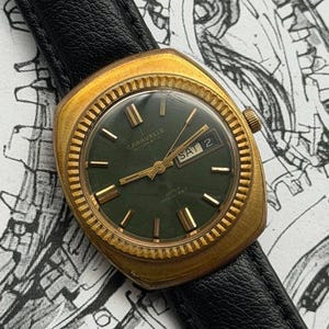 Op de afbeelding: Een vintage Caravelle automatische horloge met een goudkleurige kast en een donkergroene wijzerplaat. Het horloge heeft goudkleurige wijzers en markeringen, een dag-datumweergave en een zwarte leren band. Het horloge is waterbestendig.