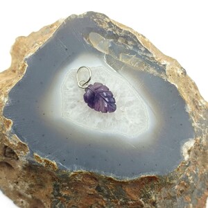 May include: A carved amethyst leaf pendant with a silver bail. The pendant is a deep purple color and is set against a gray and white agate stone. The agate stone has a brown and tan outer edge.