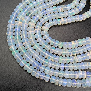 Puede incluir: Cadenas de cuentas opalescentes con un brillo suave y nacarado. Las cuentas son una mezcla de blanco y azul pálido, con toques iridiscentes. Están ensartadas, creando una forma fluida y orgánica. Cada cuenta mide aproximadamente 0,6 cm de diámetro.