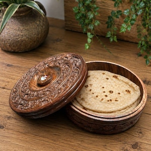 Puede incluir: Un recipiente redondo de madera marrón oscuro con una tapa tallada con motivos florales, parcialmente abierto para revelar una pila de pan plano en su interior. La tapa presenta intrincados relieves y un pequeño pomo central con una etiqueta OK.