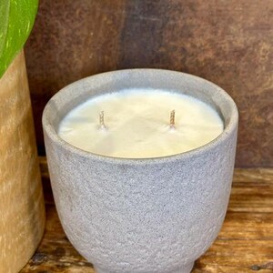 May include: A double-wick candle in a textured, light gray ceramic vessel with a tapered base. The wax is smooth, off-white, and fills the container to the brim. The two wicks are centered and upright, protruding from the surface of the wax.