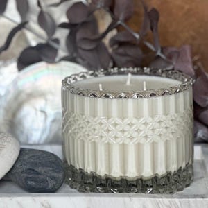 Puede incluir: Una vela decorativa en un tarro de cristal transparente con borde plateado. La vela tiene tres mechas y cera blanca. El tarro está sobre una bandeja de mármol con dos piedras y una concha en el fondo.