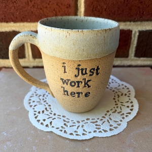 May include: A speckled ceramic mug with a light tan base and a cream-colored rim. The mug features a curved handle and the text 'i just work here' stamped in black lettering on the front. The exterior has a textured, earthy appearance.