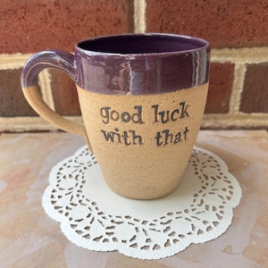 Può includere: Tazza in ceramica fatta a mano con esterno beige e interno e manico viola scuro. La tazza presenta la frase "good luck with that" in lettere nere. Poggia su un centrino di carta bianca traforata.