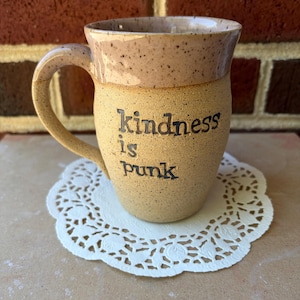 May include: A speckled ceramic mug with a light tan base and a slightly darker rim. The text "Kindness is punk" is imprinted in black on the front of the mug. The mug features a curved handle and a textured, earthy appearance throughout the surface.