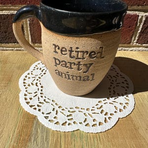 May include: A speckled tan ceramic mug with a dark blue glazed rim and handle. The front of the mug features the embossed text "retired party animal" in a dark font. The mug has a rounded body and a sturdy handle on the left side.