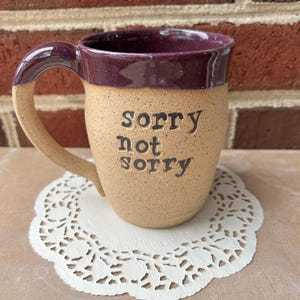 May include: A tan ceramic mug with a purple interior and handle, featuring the phrase "Sorry not sorry" in black lettering. The mug is placed on a white, lacy paper doily. The mug has a speckled texture.