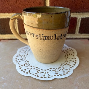 May include: A speckled beige ceramic mug with a dark olive green glazed rim and handle. The word "overstimulated" is embossed in black lowercase letters across the front of the mug. The mug has a sturdy handle and a tapered base design.