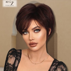 May include: A woman with blue eyes and dark red short hair. The hair is styled in a short, layered pixie cut. The image includes the text "wig" and "THALEYA" in the upper left corner. She is wearing a black top and a gold necklace.